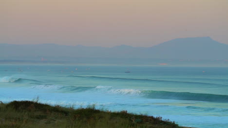 Cinematic-pan-stunning-early-morning-huge-wave-crashing-swell-surf-close-up-with-sailboats-Hossegor-Seignosse-France-pink-purple-orange-sunrise-sunset-on-beach-mountain-shoreline