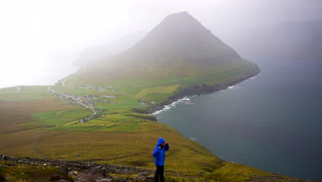 Man-wearing-a-blue-jacket-covers-himself-up-and-looks-at-Malinsfjall-Faroese-landscape,-cold-weather,-wide