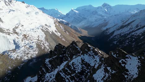 Aerial-snowy-winter-mountain-range-ridgeline-with-steep-valleys-below-on-sunny-day,-flying-around-peaks