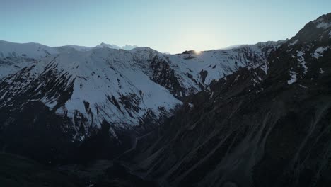 Aerial-snowy-winter-mountain-scenery-with-steep-valleys,-sun-dissapearing-behind-ridgeline
