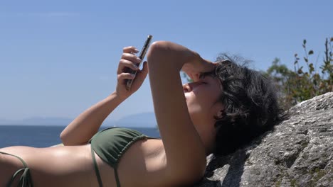 Brunette-girl-in-bikini-sunbathing-and-using-smartphone-looking-at-camera