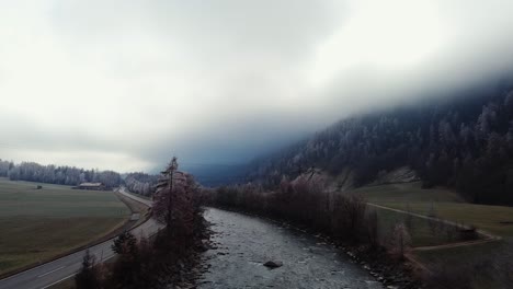 Flying-over-river-in-moody-foggy-rural-aerial-scenery,-fields-with-farm-houses-on-cloudy-day-Ötztal,-Austria