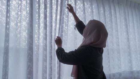Muslimische-Asiatische-Indonesische-Frau-Im-Hijab,-Die-An-Bewölkten-Tagen-Fenstervorhänge-öffnet