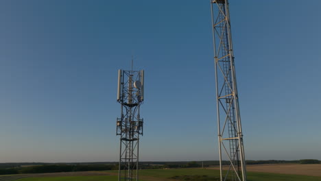 Two-cellular-towers-in-the-middle-of-farmland-with-sky-background,-aerial-orbital-closeup