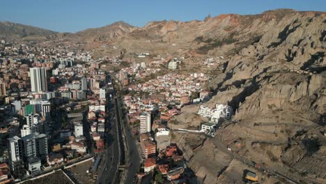 Aerial-drone-shot-over-buildings-and-houses-alongside-two-way-main-road-at-the-foothills-of-a-mountain-in-La-Paz,-Bolivia-on-a-sunny-day