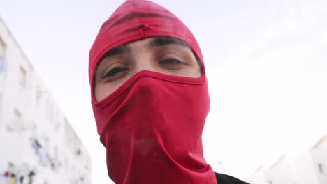 A-handsome-young-boy-man-in-a-red-full-face-mask-balaclava-exhales-steam-from-a-vape-pen-into-the-camera