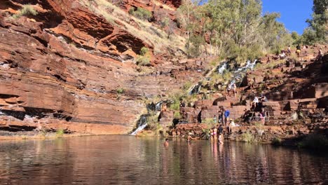 Water-pool,-red-rocks,-cliffs-and-sunbathing-tourists-at-the-Fortescue-Falls-in-Karijini,-Western-Australia