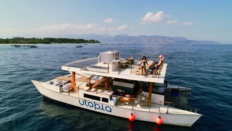 Young-Couple-Travelers-Enjoy-Rooftop-View-of-Gili-Coastline-From-Luxury-Catamaran-Yacht-Cruising-in-Blue-Sea---Aerial-Parallax-View
