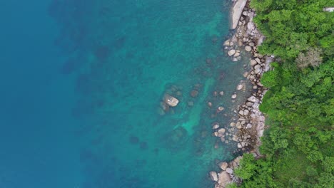 Tropical-Paradise:-A-stunning-vertical-shot-descending-drone-flight-on-a-stone-surrounded-by-water-a-stone-coast,-next-to-green-trees-and-blue-water-of-Phuket-island-in-4K