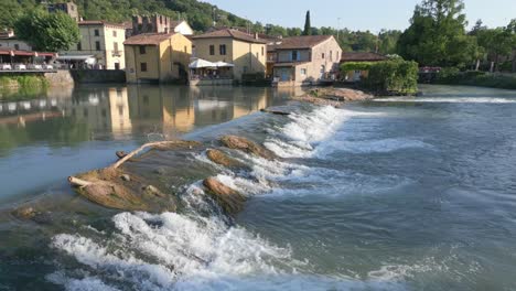 Water-flows-over-smooth-wide-shallow-waterfall-in-Borghetto-Verona-Italy,-aerial-dolly