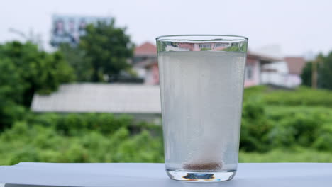Bubbly-water-in-a-glass-caused-by-dilution-of-an-antacid-or-sodium-bicarbonate-on-a-backdrop-of-houses-and-green-vegetation-out-of-focus