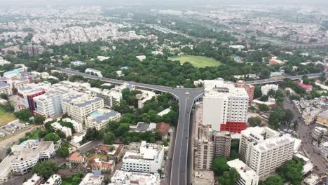 Aerial-view-of-Rajkot-city,-a-lot-of-traffic-is-going-over-the-fly-overbridge-and-tall-highrise-buildings-show-all-around-it-and-there-are-also-big-trees-and-gardens-in-the-middle-of-the-city