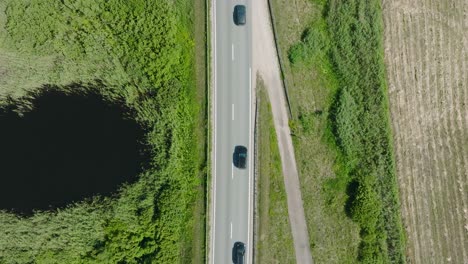 Aerial-establishing-shot-of-a-rural-landscape,-countryside-road-with-trucks-and-cars-moving,-lush-green-agricultural-crop-fields,-sunny-summer-day,-wide-descending-birdseye-drone-shot