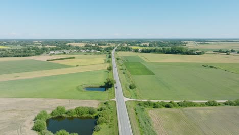 Aerial-establishing-shot-of-a-rural-landscape,-countryside-road-with-trucks-and-cars-moving,-lush-green-agricultural-crop-fields,-sunny-summer-day,-wide-drone-shot-moving-forward,-tilt-down