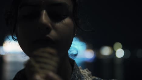 Young-beautiful-Indian-woman-eats-ice-cream-and-brushes-her-hair-from-her-forehead,-city-lights-in-the-background