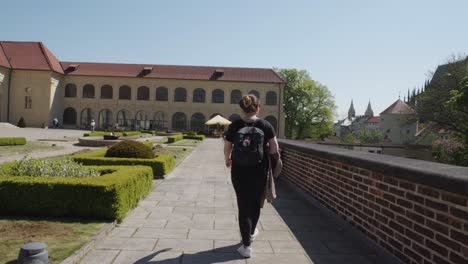 Mujer-Adulta-Caminando-Hacia-El-Edificio-De-La-Escuela-De-Equitación-Viendo-La-Torre-Del-Castillo-De-Praga-Al-Fondo-En-La-República-Checa