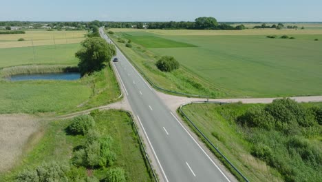 Aerial-establishing-shot-of-a-rural-landscape,-countryside-road-with-trucks-and-cars-moving,-lush-green-agricultural-crop-fields,-sunny-summer-day,-wide-drone-shot