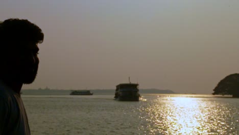 Ein-Indischer-Mann-Steht-Am-Ufer-Des-Sees-Vor-Der-Kamera,-Sonnenuntergang,-Hausboote-Im-Hintergrund,-Silhouette