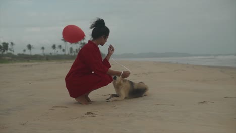 Filmaufnahme-Eines-Indischen-Models,-Das-Ein-Rotes-Kleid-Und-Einen-Roten-Luftballon-Trägt-Und-An-Einem-Windigen-Tag-Einen-Wilden-Hund-In-Goa,-Indien,-An-Einem-Sandstrand-Streichelt