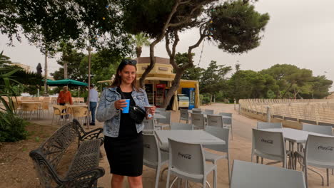 Foto-De-Una-Joven-Trayendo-Bebidas-En-Una-Mesa-En-Un-Café-Al-Aire-Libre-Junto-A-Un-Popular-Lugar-Turístico-En-Malta-En-Un-Día-Nublado
