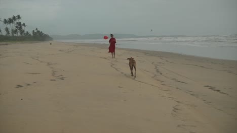 Toma-Cinematográfica-De-Una-Modelo-India-De-Moda-Corriendo-En-Una-Playa-De-Arena-Con-Un-Vestido-Rojo-Y-Un-Globo-Rojo-En-La-Mano-Mientras-Un-Perro-Camina-Detrás-De-Ella-En-Goa,-India.