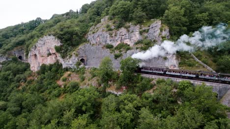 Drone-tracking-of-a-steam-train-on-the-tracks,-passing-through-a-tunnel-on-its-way-to-the-station-at-Martel,-a-village-in-the-Lot-region-of-France