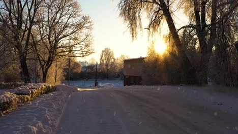 Icy-road-and-sun-shining-over-rural-village-while-snowing,-dolly-forward