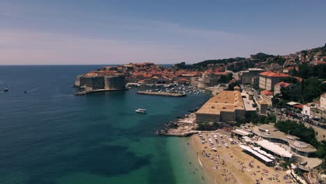 Aerial-drone-flying-over-Banje-Beach-and-aqua-blue-waters-towards-the-historic-walled-city-of-Dubrovnik,-Croatia
