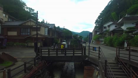 Die-Drohnenansicht-Schwebt-über-Dem-Wasserlauf-Eines-Japanischen-Dorfes-Und-Bewegt-Sich-Auf-Eine-Brücke-Zu,-An-Der-Zwei-Frauen-Mit-Regenschirmen-Bei-Mildem-Regen-Vorbeikommen