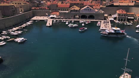 Aerial-drone-flying-over-the-port-of-Dubrovnik,-Croatia-filled-with-fishing-boats-and-ferrys-continuing-over-the-terra-cotta-rooftops-of-the-walled-city-and-over-cathedrals-and-palaces