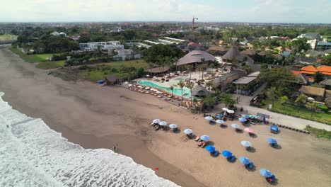 Aerial-Approaching-Mari-Beach-Club-Bali-from-Batu-Belig-Beach,-Drone-Descending-Towards-Plunge-Pool-Lounge-Area-Surrounded-with-Sunbeds-and-Daybeds,-White-Foamy-Waves-Rolling-Over-Soft-Sand,-Indonesia