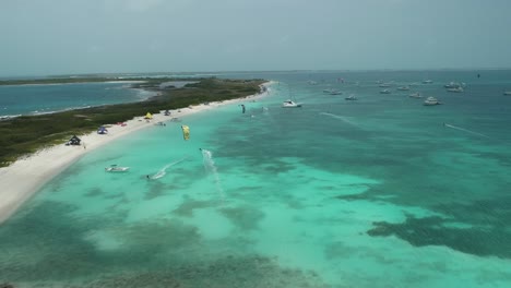 Kite-surfers-ride-on-clear-blue-ocean-water-above-reefs,-crasky-los-roques,-aerial-dolly