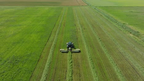 Aerial-establishing-view-of-a-tractor-mowing-a-fresh-green-grass-field,-a-farmer-in-a-modern-tractor-preparing-food-for-farm-animals,-sunny-summer-day,-wide-drone-shot-moving-forward,-tilt-down