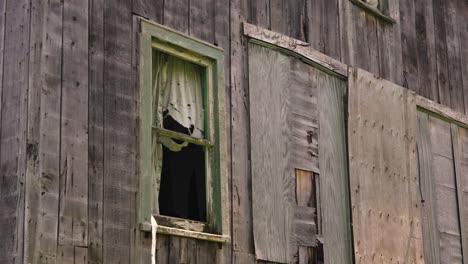 Wind-Weht-Zerrissener-Vorhang-Aus-Zerbrochenem-Holzfenster,-Von-Termiten-Befallenes-Haus,-Mit-Brettern-Vernagelte-Tür