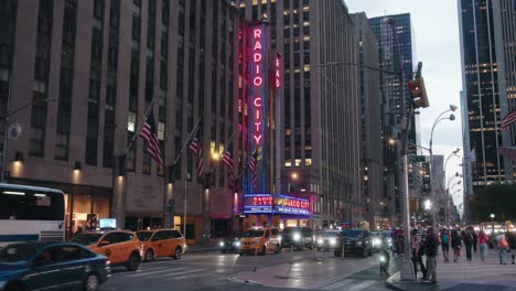 Radio-City-Music-hall-neon-sign,-full-profile-side-view,-mopeds-and-cars-drive-by-with-lights-on