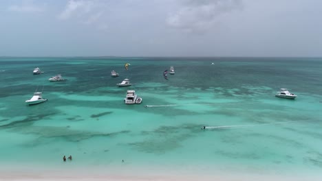 Two-kite-surfers-fly-coasting-over-stunning-blue-ocean-water-past-yachts-in-los-roques,-aerial-tracking
