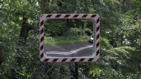 A-convex-road-safety-mirror-set-in-woodland-at-a-road-junction-as-vehicles-pass-by,-Worcestershire,-England