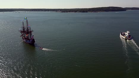 An-aerial-view-of-a-17th-century-wooden-ship-and-a-Greenport-North-Ferry,-both-heading-out-on-Greenport-Harbor-on-Long-Island-on-a-sunny-day