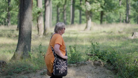 Panning-shot-of-an-older-fat-Woman-with-grey-Hair-wearing-orange-T-shirt-and-black-bag-walking-through-the-green-Forest,-holding-wooden-basket-in-slow-motion-during-a-day