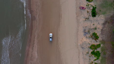 Aerial-top-down-follow-tracking-shot-of-a-car-moving-on-a-sand-beach-near-the-water-during-dusk
