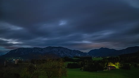 Timelapse-of-impressive-mountain-landscape-in-Austria-with-fast-moving-clouds-and-view-of-the-imposing-mountains-and-a-house,-which-is-illuminated-and-sets-warm-light-accents-in-the-evening