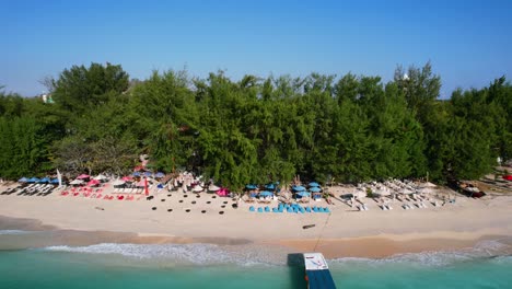 Sand-Tourist-Beach-on-Gili-Trawangan-Island-with-Empty-Umbrellas-and-Deck-Chairs-as-Turquoise-Waves-Crash-in-the-Morning---Aerial-Parallel-Dolly