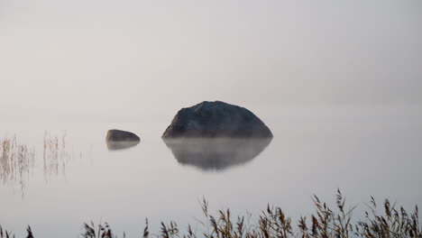 Peaceful-still-atmosphere-lake-water-with-stone-in-middle,-hazy-fog-moving