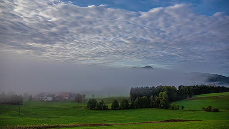 Timelapse-of-austrian-landscape-with-picturesque-meadow-with-colorful-buildings-in-a-layer-of-fog-in-the-early-morning-with-a-view-of-the-hills-and-mountains-with-fast-moving-clouds