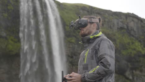 Man-controls-an-FPV-drone-for-aerial-photography-and-video-with-a-remote-control-and-goggles,-behind-a-waterfall-in-Iceland