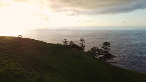 Luftaufnahme-Eines-Dollys-über-Der-Friedlichen-Küstenlandschaft-Mit-Sanft-Abfallenden-Hügeln-Und-Einem-Weiten-Blick-Auf-Den-Horizont-Mit-Spiegelndem-Meer-Auf-Der-Insel-Norfolk-Zur-Goldenen-Stunde