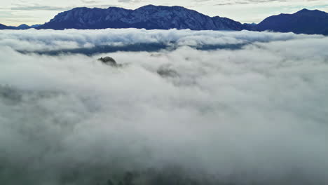 Aerial-shot-of-hilly-landscape-covered-by-thick-layer-of-clouds-with-visible-snowcapped-mountain-in-the-background