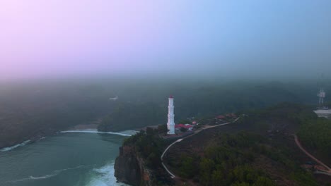 Aerial-view-of-white-lighthouse-on-the-hilly-shoreline-in-the-foggy-evening-sky---Baron-beach,-Yogyakarta,-Indonesia