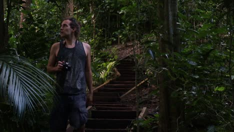 Static-shot-of-a-tourist-with-camera-in-hand-in-the-middle-of-a-densely-overgrown-forest-while-walking-down-a-wooden-staircase-and-observing-the-beauty-of-nature-on-his-journey-through-the-jungle
