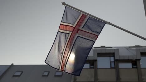 Icelandic-flag-waving-in-the-wind,-with-buildings-behind-it,-in-slow-motion,-Iceland